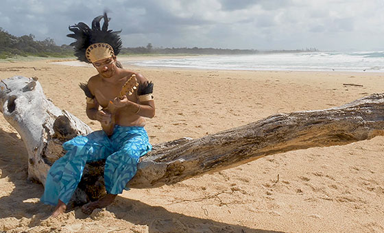 A man wearing flowy pants and a headdress sits on a beached tree and plays a small guitar.