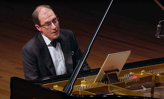 A man wearing glasses and a formal suit sits in front of an open-topped piano while he sways to his playing.