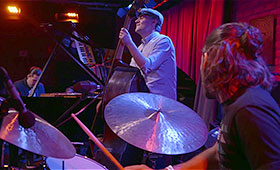 A drummer and bass player perform in the foreground as Rodriguez plays the piano in the background.