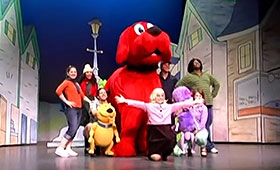 Seven actors and two small dog puppets stand around a big red dog puppet in the middle of a stage set.