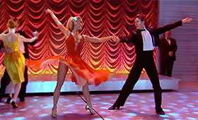 A woman in a twirly skirt and a man in a tuxedo dance together with their arms extended.