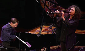 Fred Hersch looks down at the keys while he plays the piano as Anat Cohen points her clarinet straightforward while playing it.