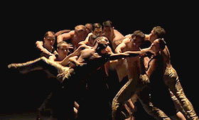 A male dancer extends his arms and legs out while his body is supported by a large group of male and female dancers.