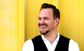 A man with a goatee and wearing a white shirt and black vest smiles at the camera in front of a metal wall.