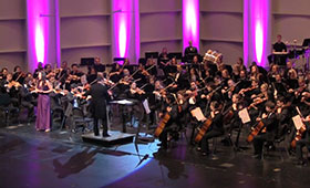 An orchestra performs on a brightly lit stage while a woman sings next to the conductor.