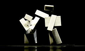 Two humanoid figures composed of boxes stand on stage. One taps the shoulder of the other.