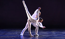 A male dancer supports a female dancer as she leans down toward the stage with a leg extended to the sky.