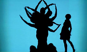 Dancers pose behind a screen to create a silhouette of a flowering plant while the silhouette of a young girl approaches it.