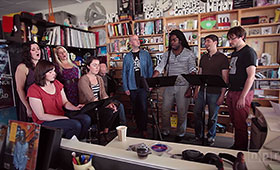 The members of Roomful of Teeth gather around three music stands and in front of various bookcases in a photo taken during an NPR Tiny Desk Concert.
