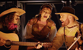 A man with a guitar, a woman, and a mad with a fiddle sing around a campfire.