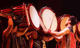 A man plays a row of large drums, each supported on the shoulders of other musicians.